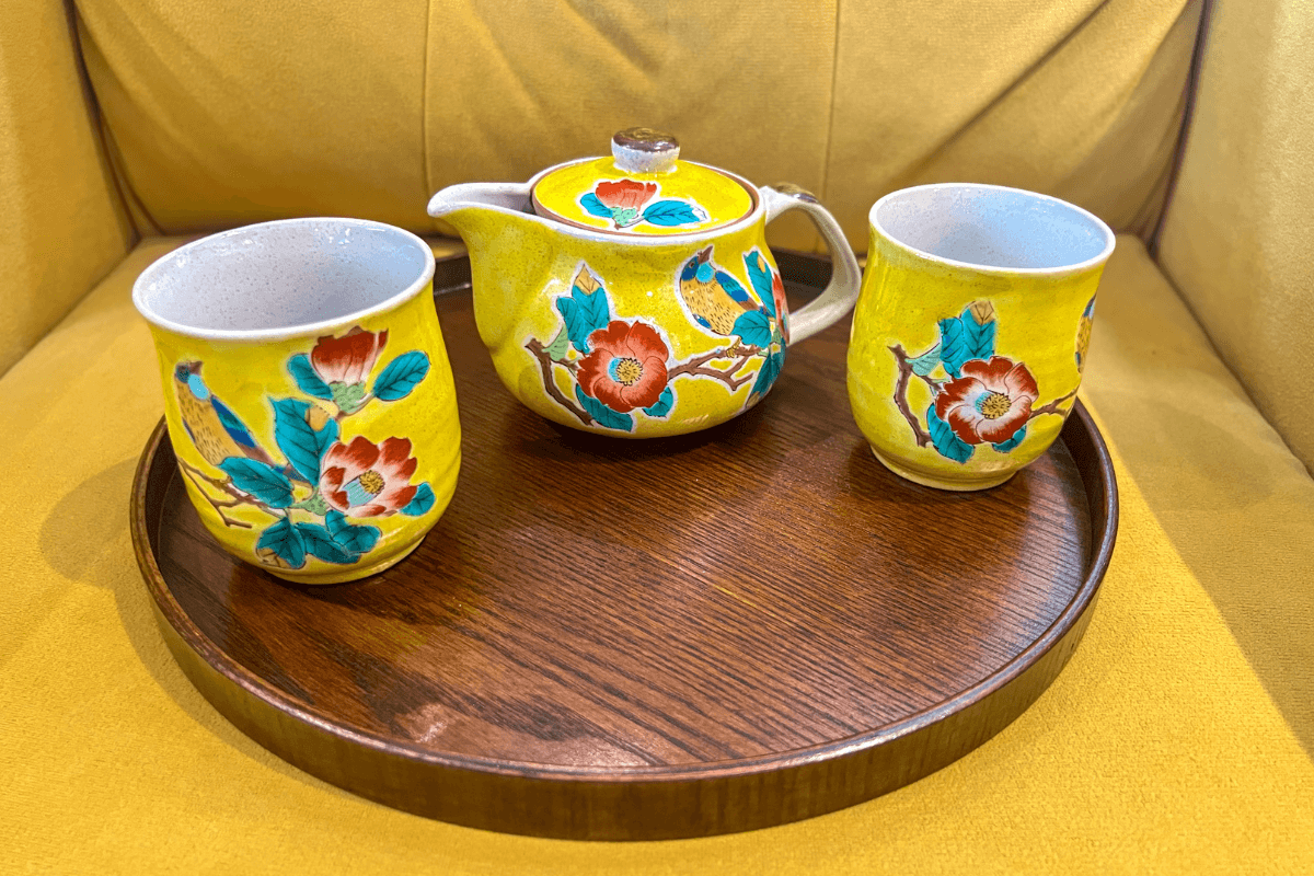 20 Memphis FINDS to See You Through the End of Summer - 4 A yellow floral teapot with a lid and two matching cups sit on a round wooden tray, placed on a yellow upholstered surface.