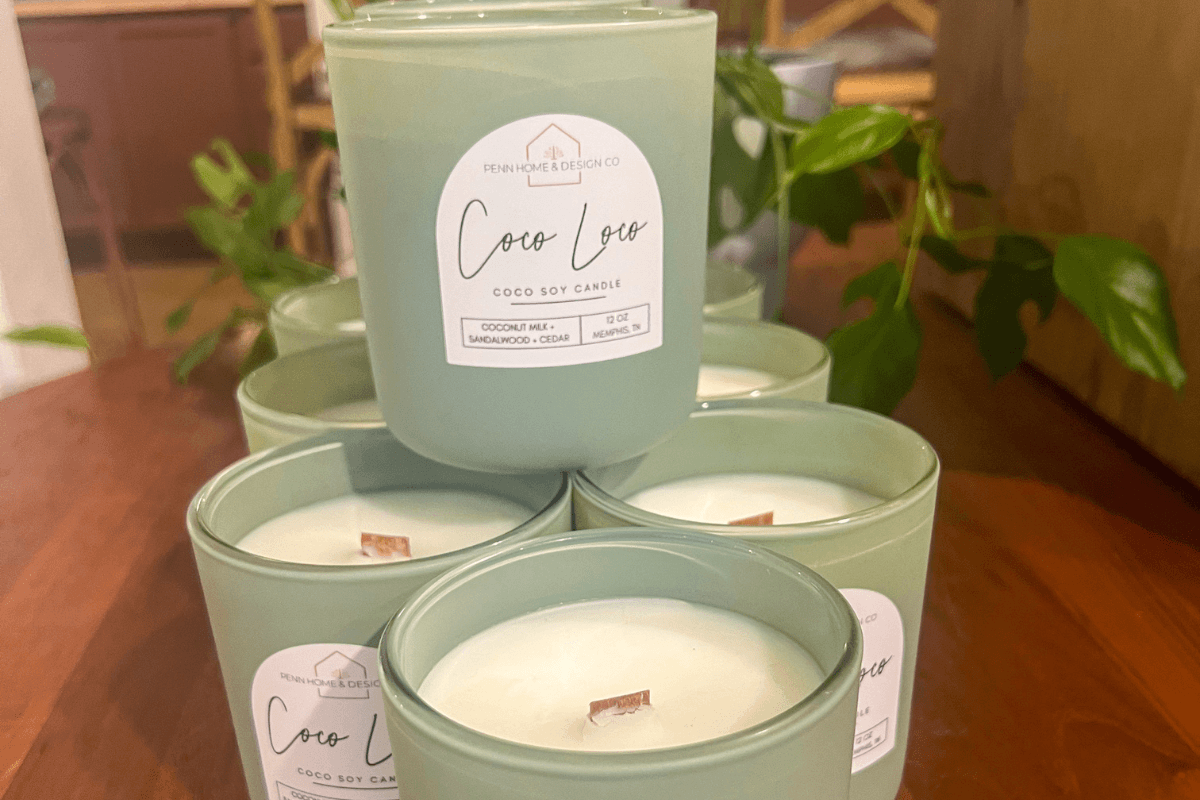 20 Memphis FINDS to See You Through the End of Summer - 21 A stack of six green glass candles labeled "Coco Luxe Coco Soy Candle" with wooden wicks, displayed on a wooden surface next to green leafy plants.
