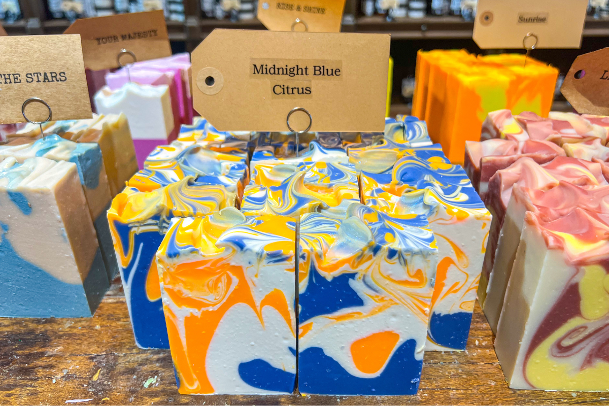 20 Memphis FINDS to See You Through the End of Summer - 17 Four bars of handmade soap labeled "Midnight Blue Citrus" with blue, yellow, orange, and white swirled patterns are displayed on a wooden surface among other colorful soaps.