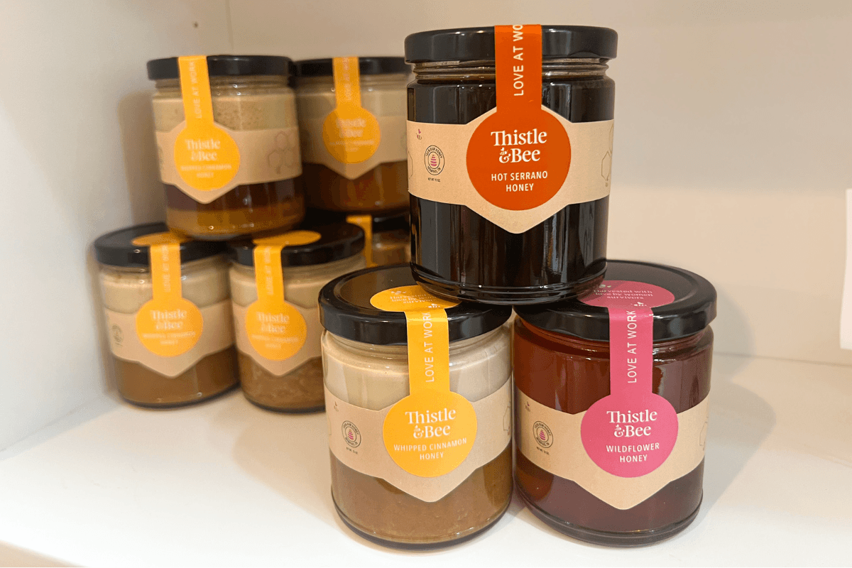 20 Memphis FINDS to See You Through the End of Summer - 14 Several jars of Thistle & Bee branded honey in different flavors are stacked on a white shelf. Labels indicate varieties like Hot Serrano, Infused Cinnamon, and Wildflower Honey.