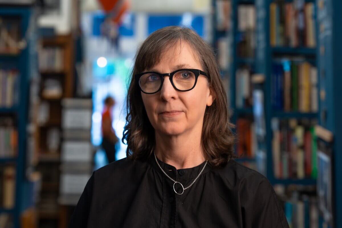 A person with shoulder-length brown hair and glasses stands in a library aisle, wearing a black shirt and two silver necklaces. Shelves of books are visible in the background.