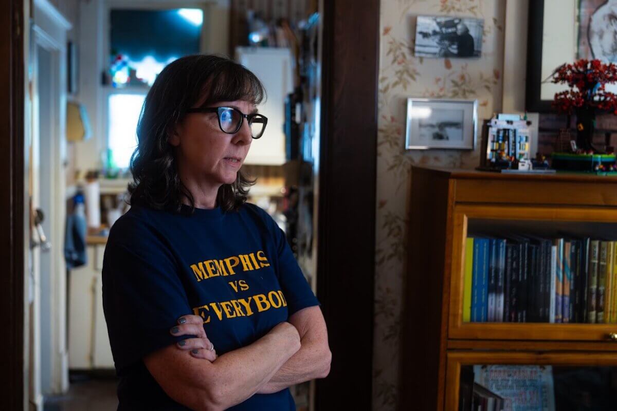 A woman with glasses stands indoors with arms crossed, wearing a "Memphis vs Everybody" shirt. Bookshelves, framed photos, and a window are visible in the background.