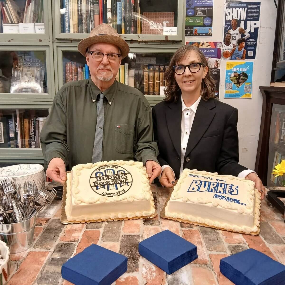 Two people in formal attire stand behind a table with two decorated cakes in a bookstore setting, with shelves of books and posters visible in the background.