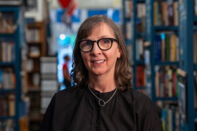 A woman with glasses and shoulder-length hair stands smiling in a library aisle, surrounded by tall shelves filled with books.