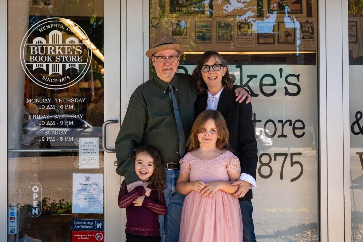 Four people, two adults and two children, stand smiling outside the entrance of Burke’s Book Store. The storefront glass displays the store name and hours of operation.