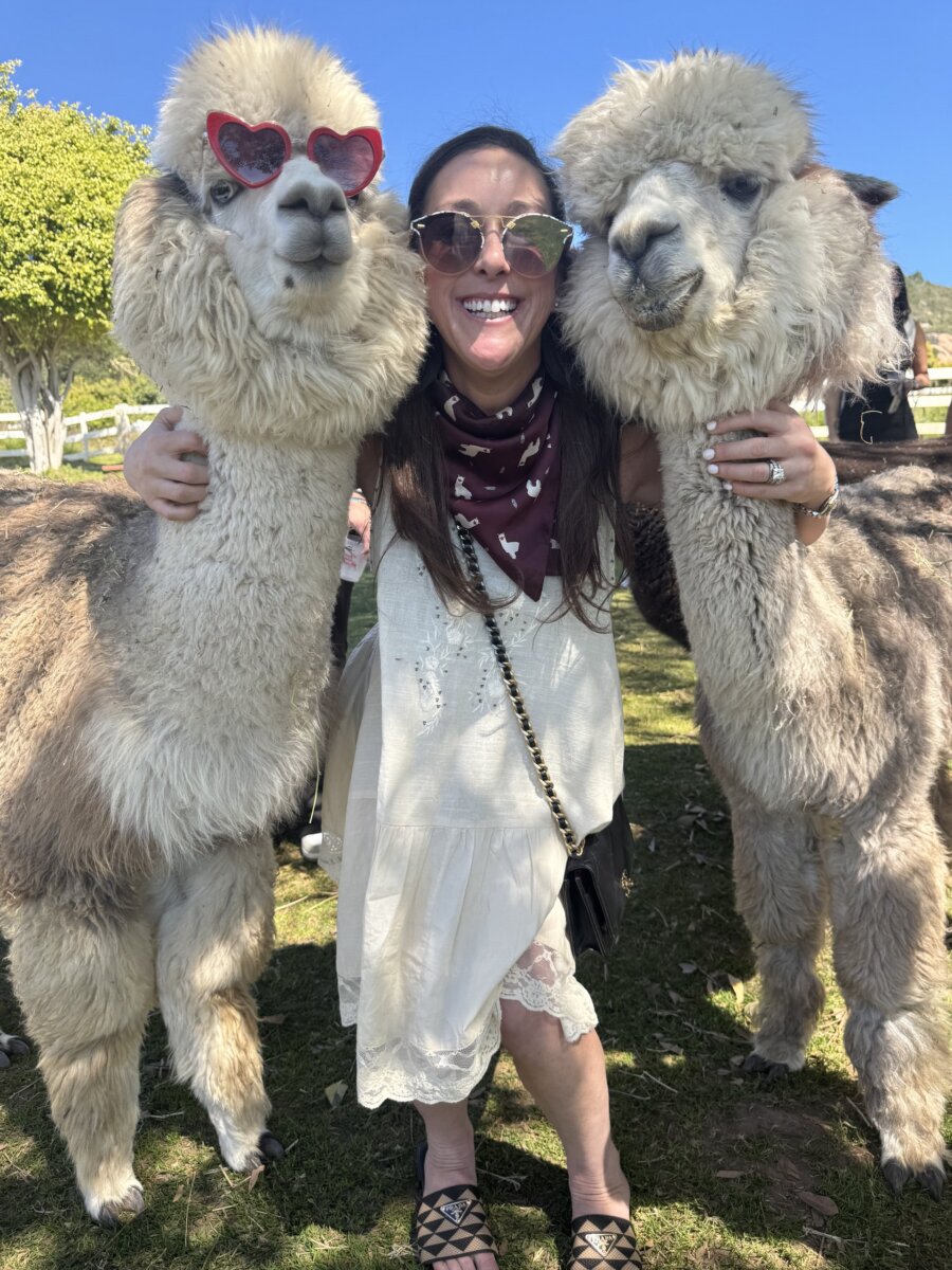She's Building Memphis's Newest Stomping Grounds - 3 Lindsey Shanmugasundaram stands outdoors between two alpacas, each wearing sunglasses. She is smiling, beaming with joy as she wraps her arms around the alpacasβ necks. Trees and a fence are visible in the background.