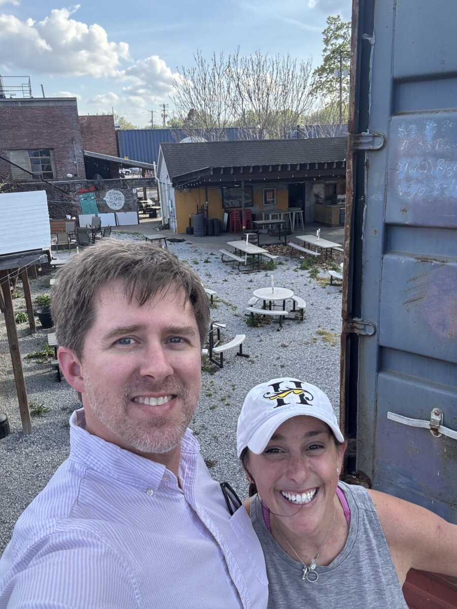 She's Building Memphis's Newest Stomping Grounds - 2 Two people, including Lindsey Shanmugasundaram, are smiling and posing for a selfie in a gravel outdoor area with picnic tables, buildings, and blue sky in the background. Their happy faces capture the cheerful atmosphere of the moment.