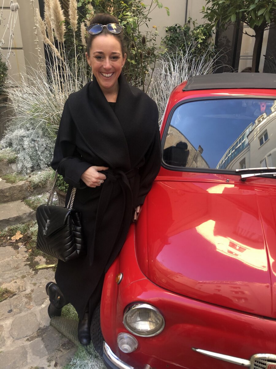 She's Building Memphis's Newest Stomping Grounds - 4 A woman in a black coat and sunglasses stands next to a vintage red car in an outdoor courtyard with plants and stone pavement.
