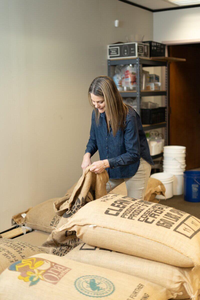 Meet the Woman Behind Muddy's Bake Shop's Next Chapter - 3 A woman in a blue shirt is opening a large burlap sack in the storage room of Muddy’s Bake Shop, with other sacks and metal shelves in the background.