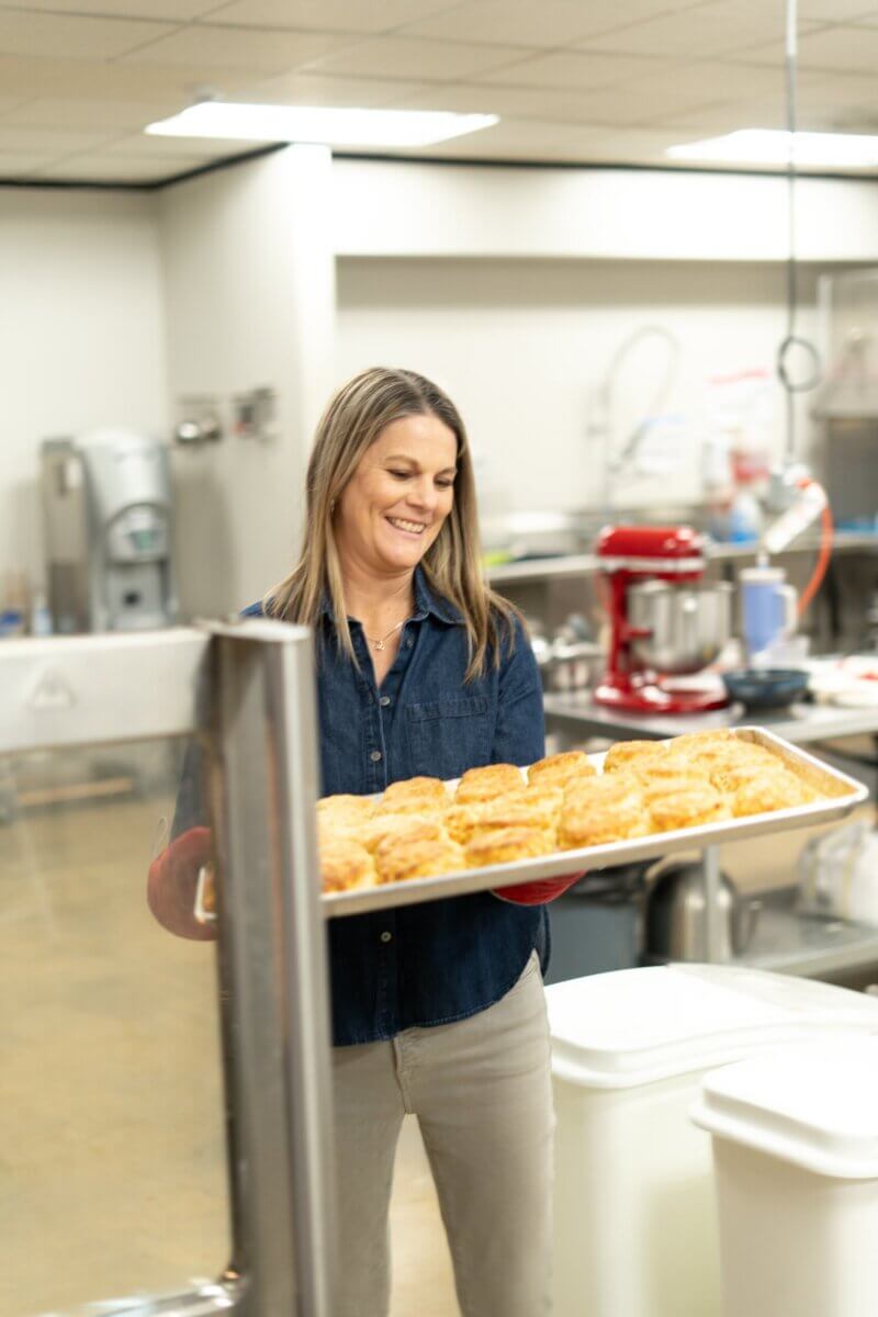 Meet the Woman Behind Muddy's Bake Shop's Next Chapter - 4 A woman in a commercial kitchen at Muddy's Bake Shop holds a large tray of freshly baked biscuits, smiling as she carries them.