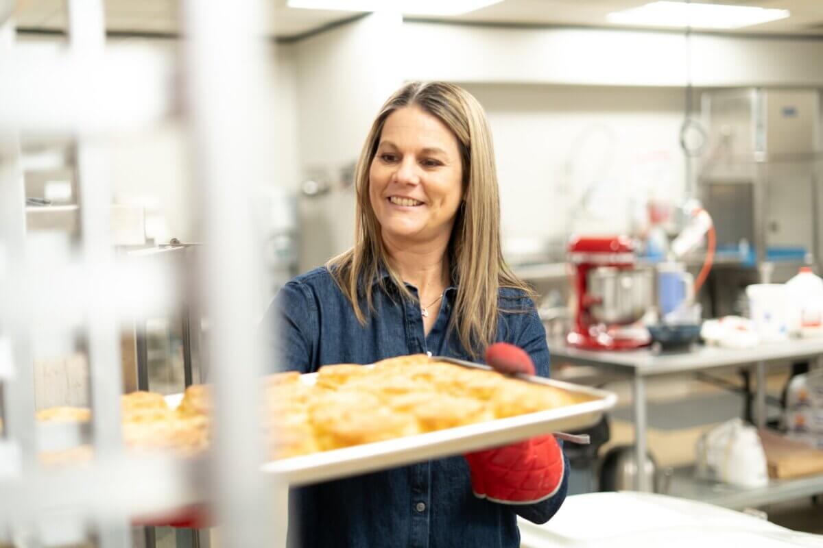 Meet the Woman Behind Muddy's Bake Shop's Next Chapter - 6 Woman wearing a denim shirt and red oven mitt holds a tray of baked goods in a commercial kitchen setting at Muddy’s Bake Shop.