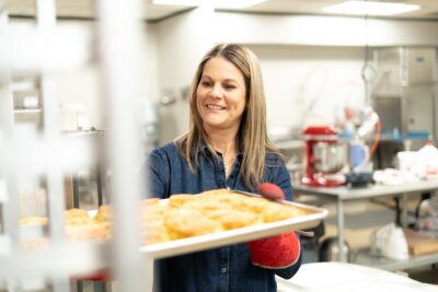 Woman wearing a denim shirt and red oven mitt holds a tray of baked goods in a commercial kitchen setting at Muddy’s Bake Shop.