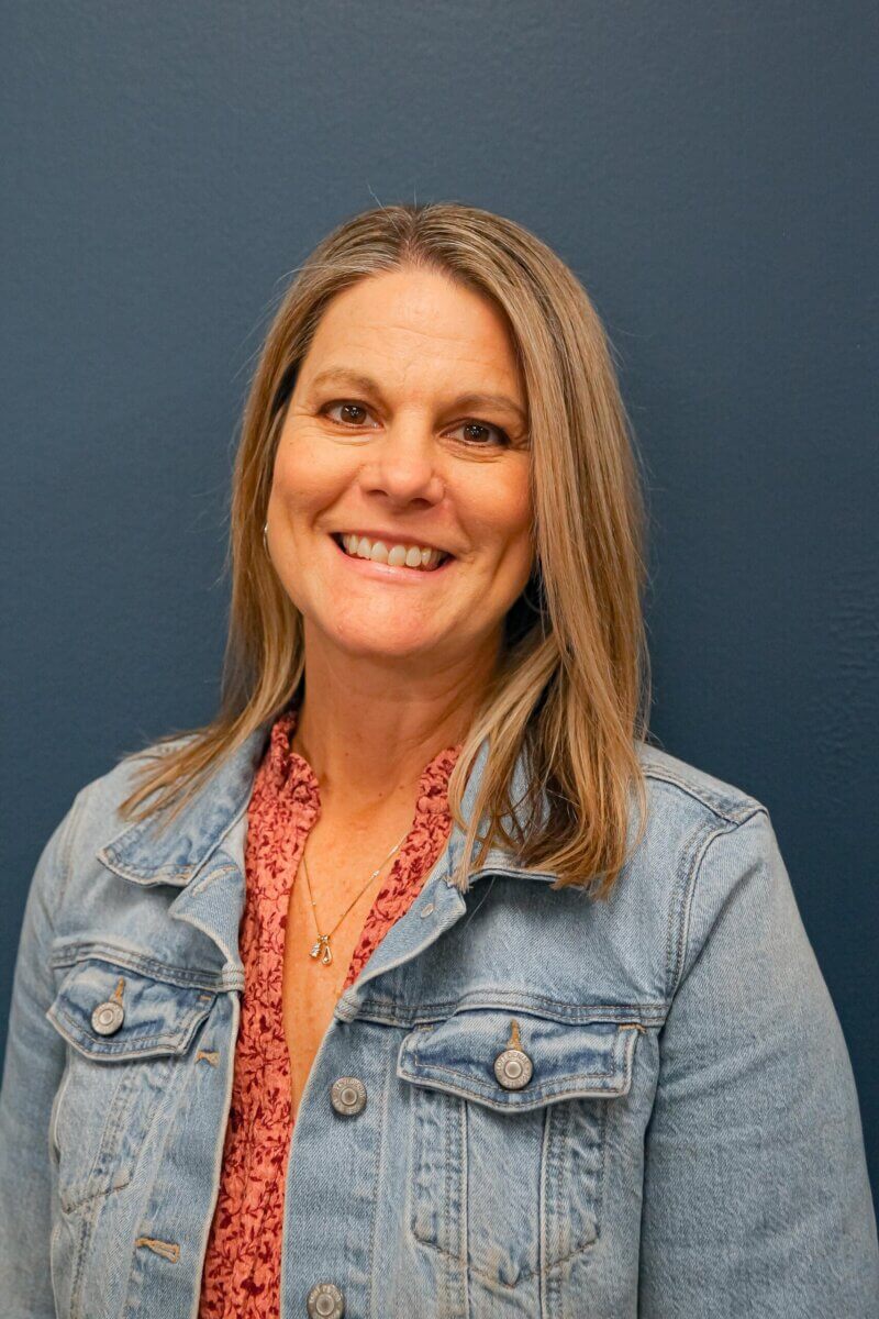 Meet the Woman Behind Muddy's Bake Shop's Next Chapter - 2 Woman with straight, shoulder-length hair wearing a denim jacket over a patterned blouse, smiling in front of a plain blue wall.