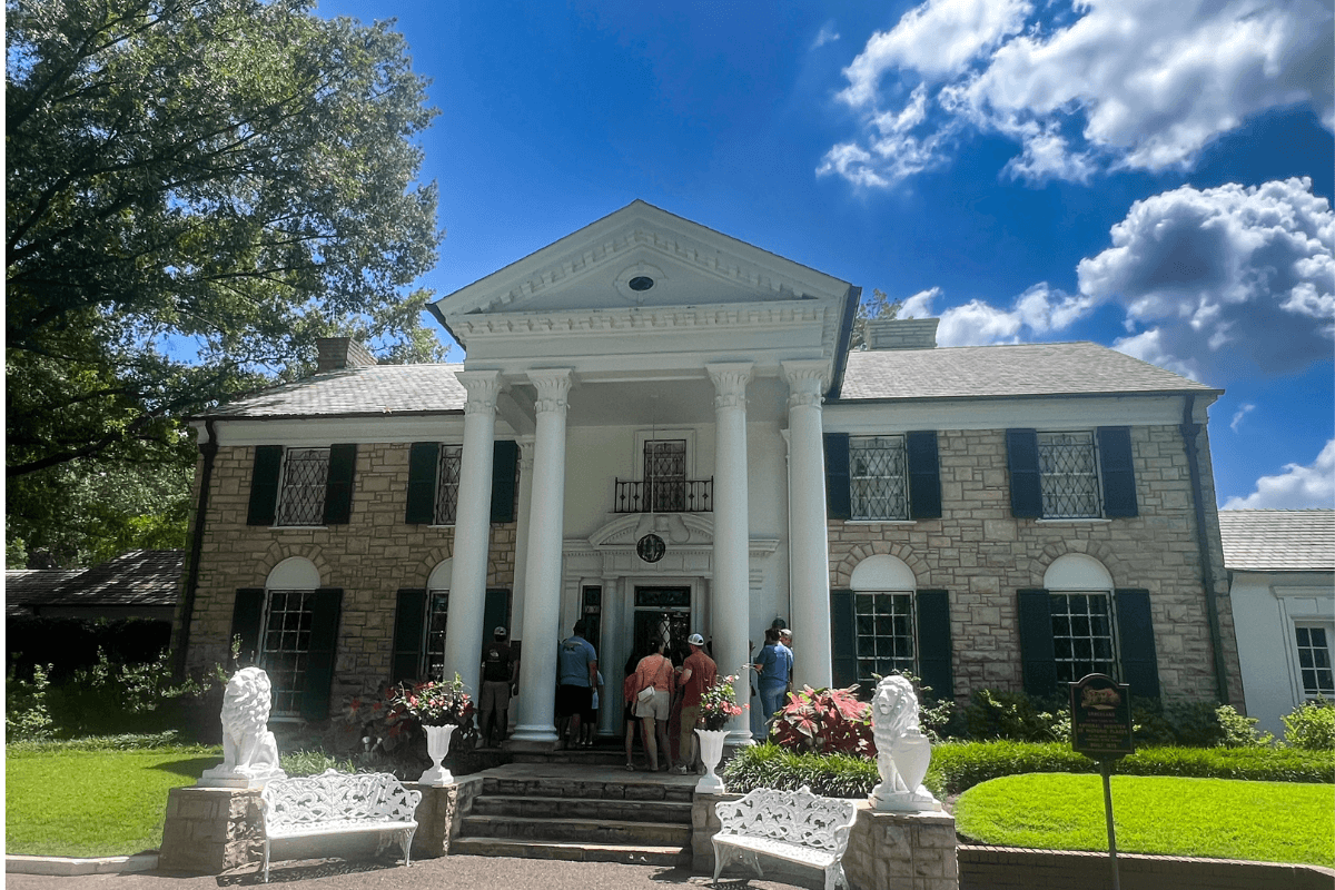 A group of people stands near the entrance of a two-story stone mansion with white columns, black shutters, and landscaped surroundings under a partly cloudy sky.