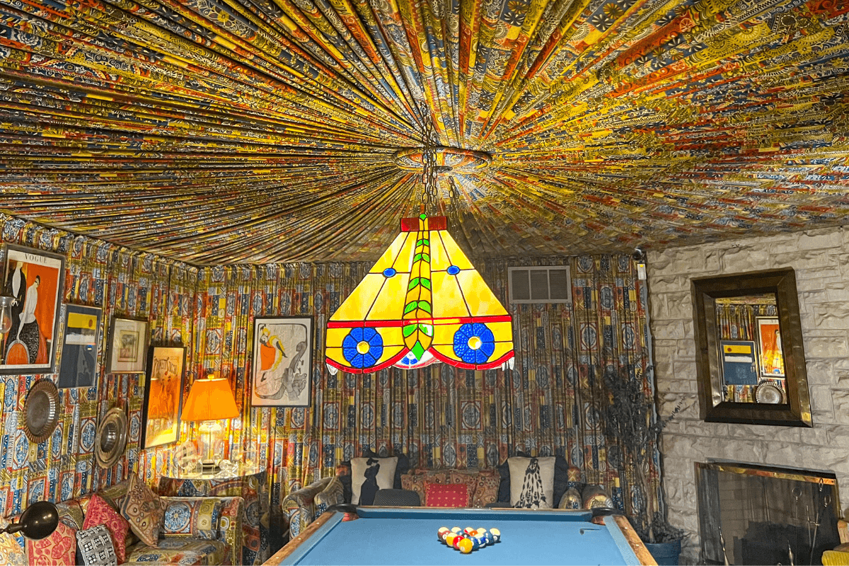 Room with patterned fabric covering walls and ceiling, stained glass lamp above a pool table, and eclectic decor including framed art and a stone fireplace.