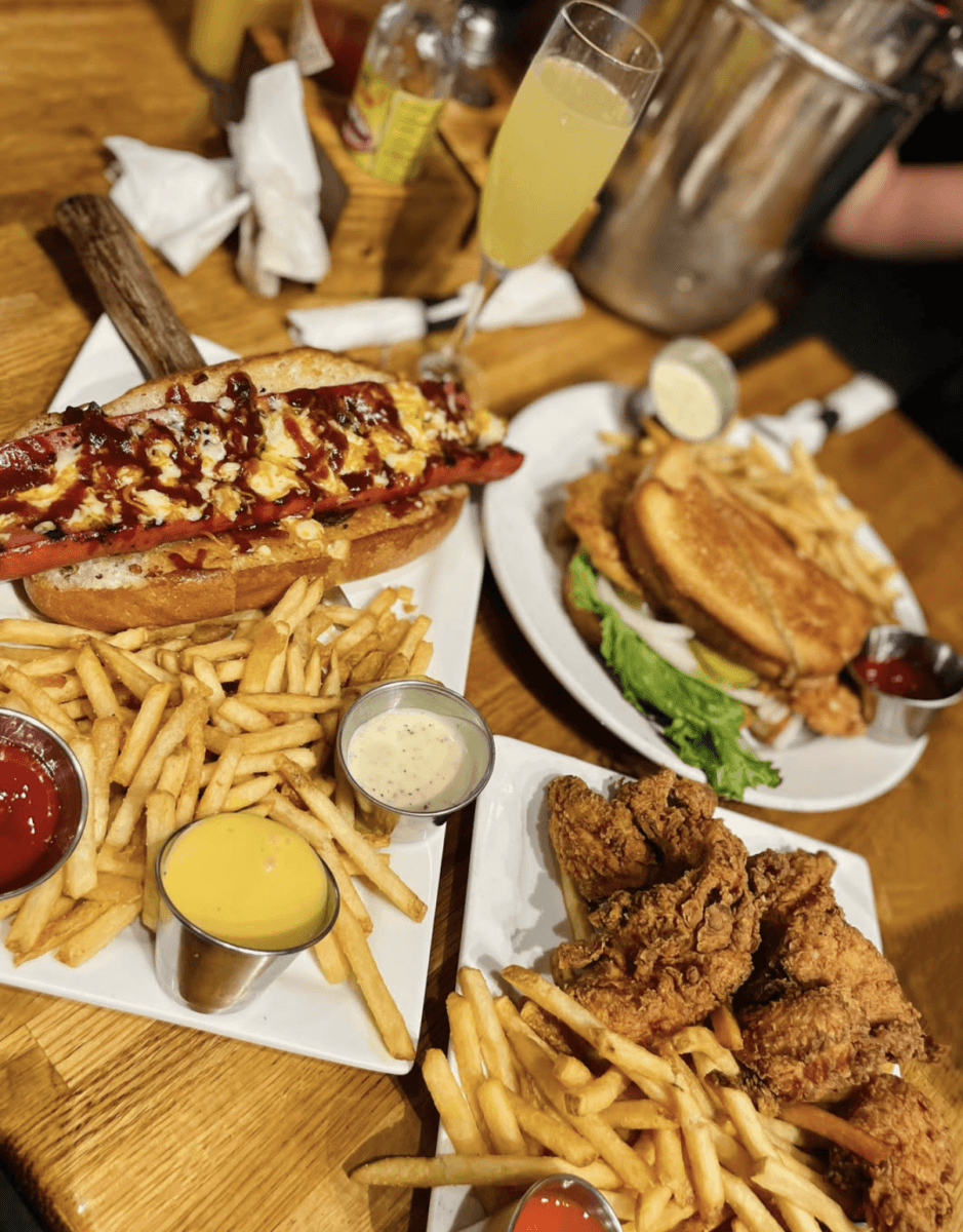 8 Memphis Hot Dogs That Go Way Beyond the Basics - 4 Three plates of food on a wooden table: a hot dog with fries, fried chicken with fries, and a sandwich with fries. Dipping sauces and a glass of orange beverage are also visible.