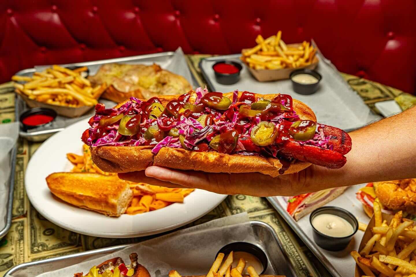 A hand holds a hot dog topped with barbecue sauce, jalapeños, and slaw. Plates of fries, sandwiches, and dipping sauces are on the table in the background.