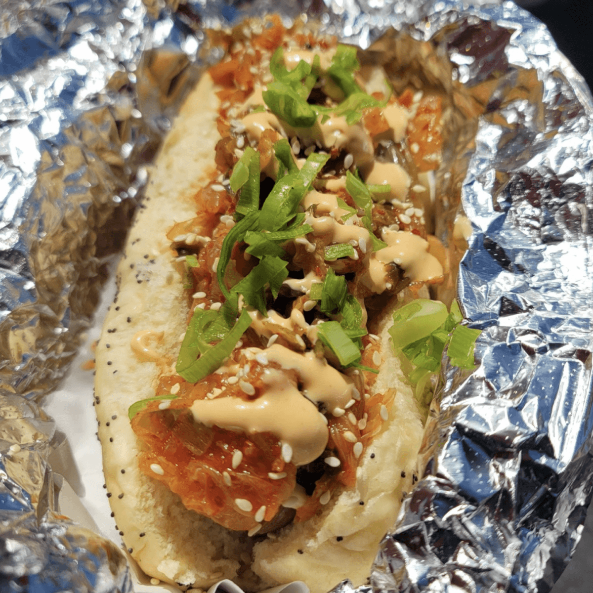 8 Memphis Hot Dogs That Go Way Beyond the Basics - 3 A hot dog topped with kimchi, green onions, sesame seeds, and a drizzle of creamy sauce, served in a poppy seed bun on foil.