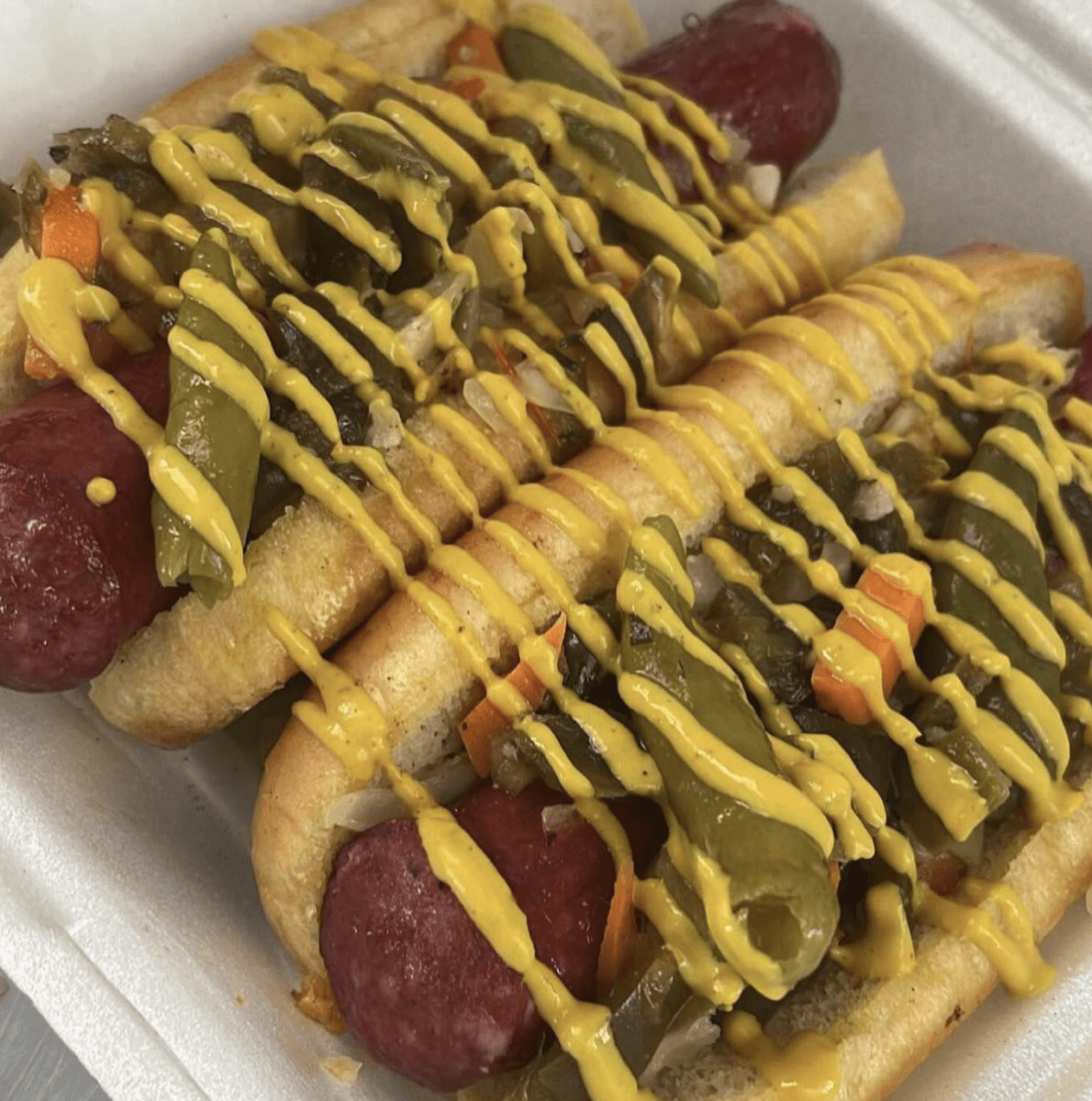 8 Memphis Hot Dogs That Go Way Beyond the Basics - 2 Two hot dogs in buns topped with chopped pickled vegetables and drizzled with yellow mustard, served in a white foam takeout container.