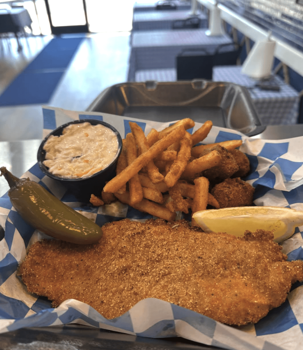 A basket with fried fish, seasoned fries, coleslaw, hush puppies, a jalapeño, and a lemon wedge on blue and white paper in a casual dining setting.
