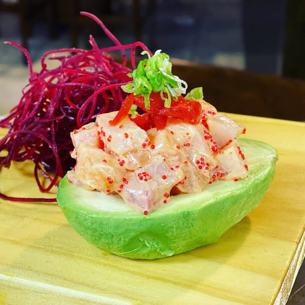 Diced fish in creamy sauce with roe served in half an avocado, garnished with scallions and red strips, next to shredded purple garnish on a wooden surface.