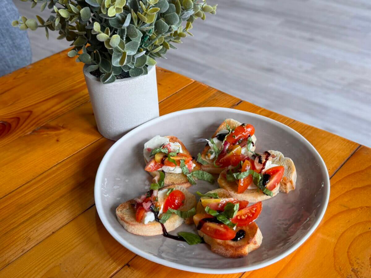 A plate of bruschetta topped with tomatoes, basil, and balsamic glaze sits on a wooden table next to a small potted plant.