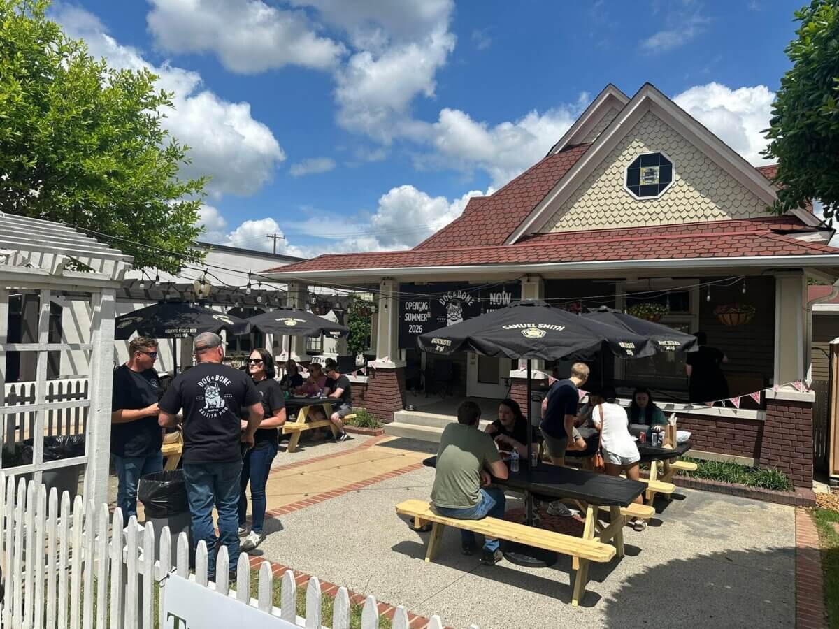 People sitting at picnic tables and standing outside a cafe on a sunny day, with umbrellas providing shade and a white picket fence in the foreground—perfectly capturing the inviting atmosphere of new restaurants coming soon to the Memphis dining scene.