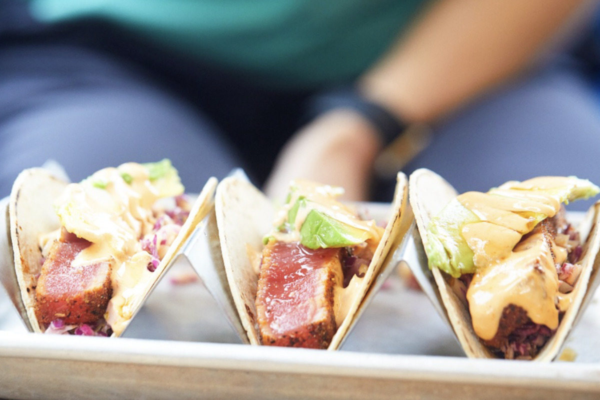 Three fish tacos on a tray, each with seared fish, sliced avocado, shredded cabbage, and a drizzle of orange sauce.