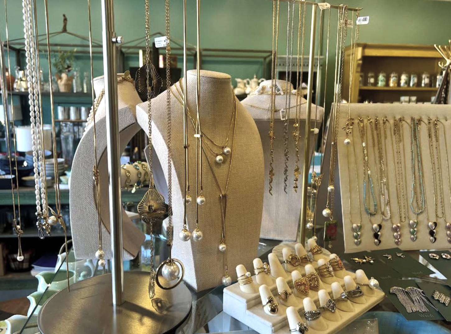 Display of assorted necklaces, rings, and earrings on racks, mannequins, and stands in a jewelry store with a green background.