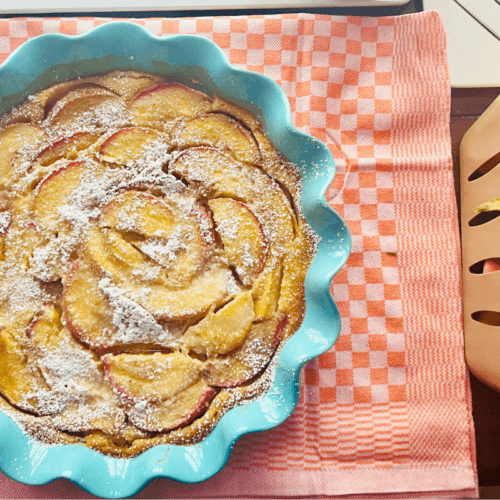 A baked peach tart dusted with powdered sugar in a blue scalloped dish sits on a pink cloth next to a basket of fresh peaches.