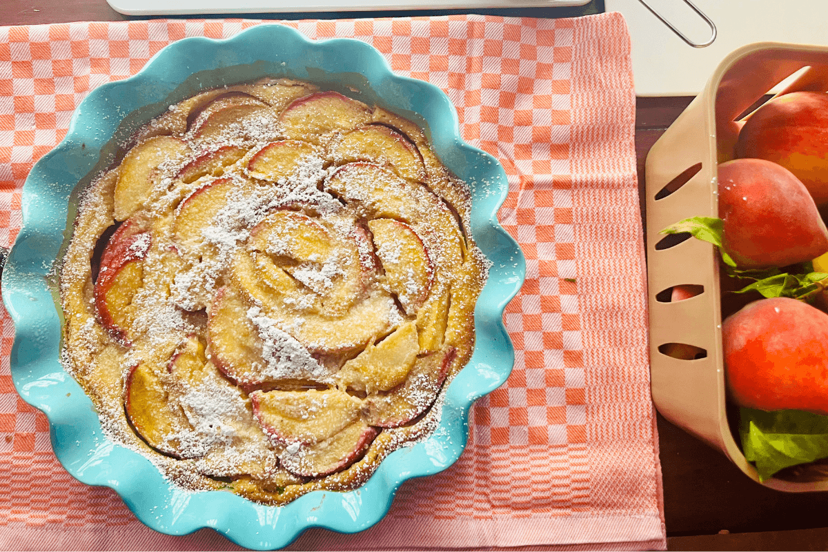 A baked peach tart dusted with powdered sugar in a blue scalloped dish sits on a pink cloth next to a basket of fresh peaches.