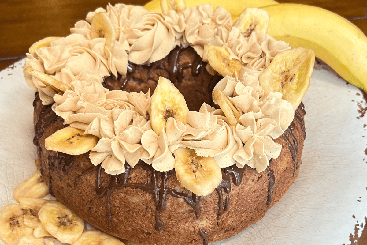 A round banana cake with chocolate drizzle, piped frosting, and banana chips on top, with whole bananas in the background.
