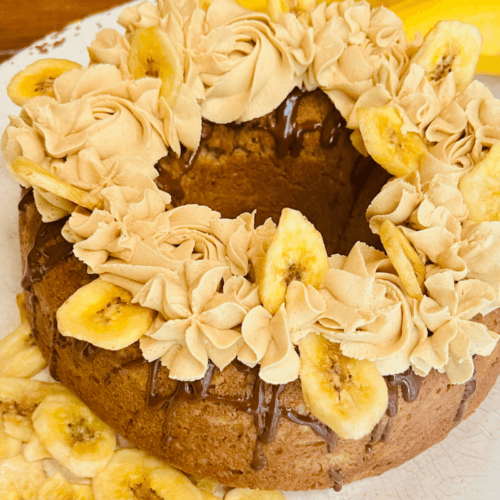 A round cake decorated with piped frosting and banana chips sits on a plate, with a whole banana in the background.