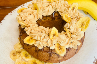A round cake decorated with piped frosting and banana chips sits on a plate, with a whole banana in the background.