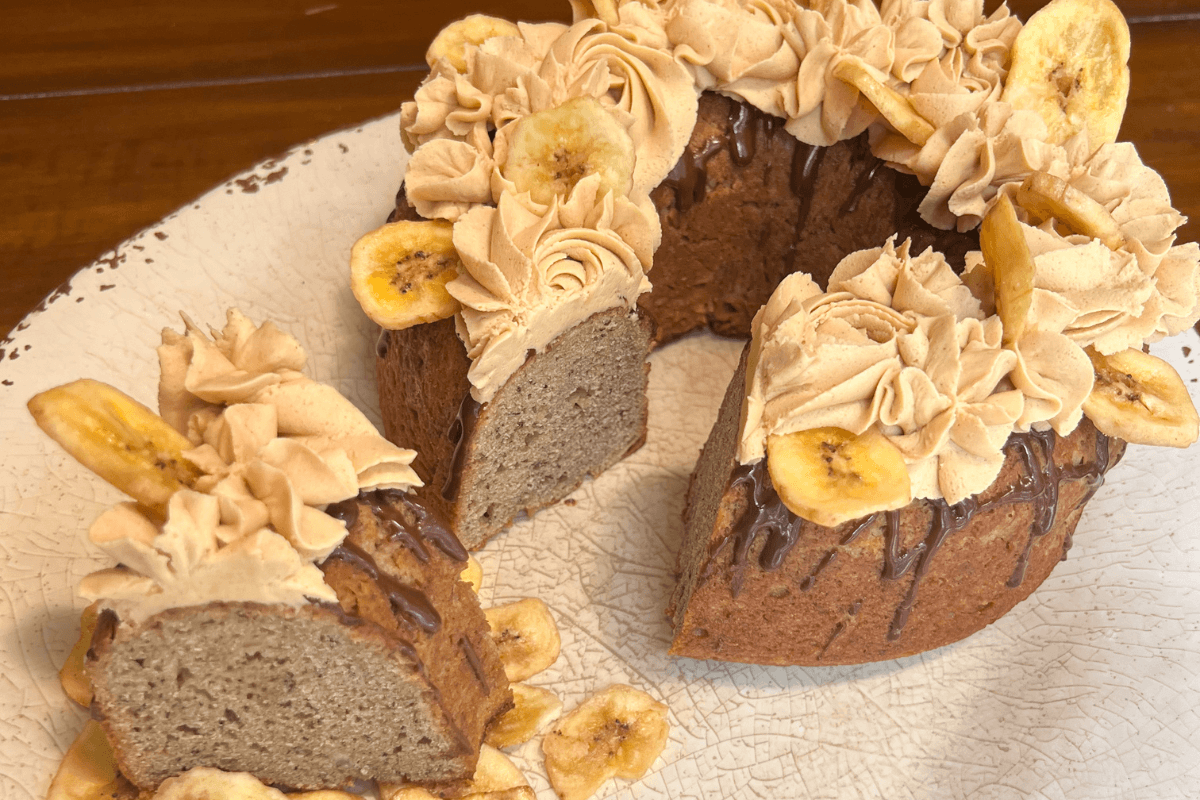 A sliced bundt cake with light brown frosting, chocolate drizzle, and banana chips on top, displayed on a white plate.