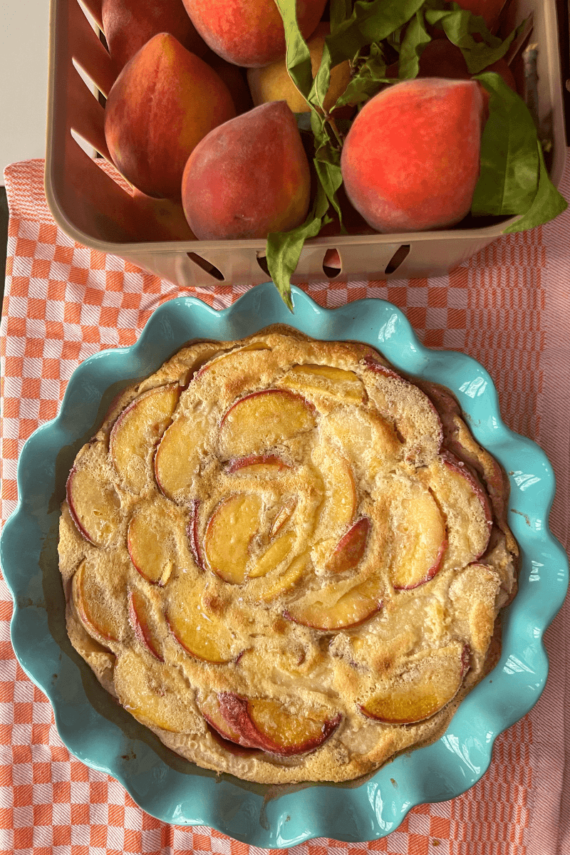 A peach tart in a blue pie dish sits on a checkered cloth next to a basket filled with whole peaches with leaves, evoking the rustic charm of a classic peach clafoutis.