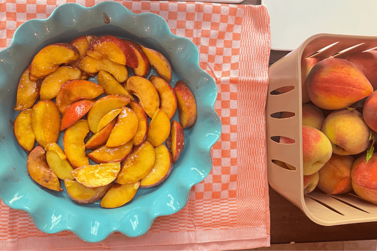 A blue pie dish filled with sliced peaches sits on a pink checkered towel next to a basket of whole peaches.