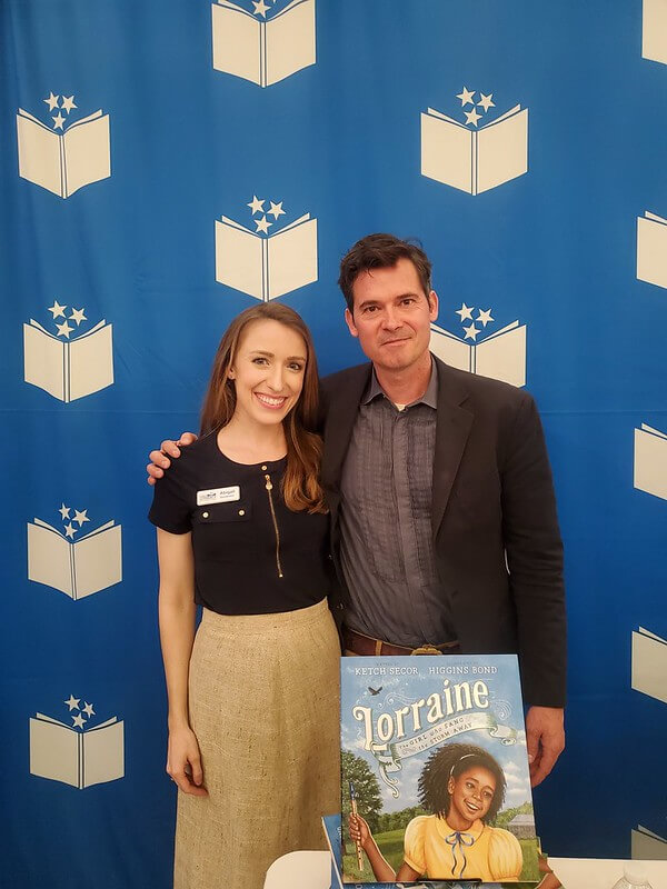 She’s Helping Put Books in the Hands of 500,000 Tennessee Kids - 11 Two people stand side by side, smiling, in front of a blue backdrop with book logos. A book titled "Lorraine" is displayed on the table in front of them.