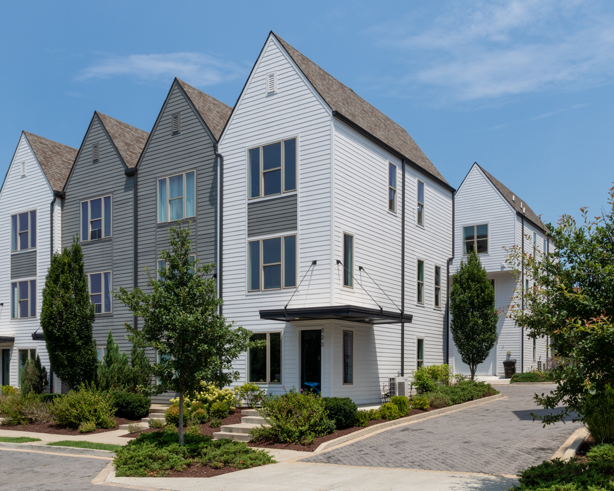 A row of modern, white three-story townhouses with peaked roofs stands along 323 East Village Ln in East Village Nashville, surrounded by green landscaping and a paved driveway under a clear blue sky.