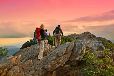 Two hikers with backpacks walk up a rocky mountain trail at sunset, the pink-orange sky and scattered clouds painting a perfect backdrop for their NC mountain road trip along the scenic Mayland Meander.