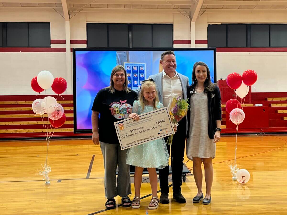 She’s Helping Put Books in the Hands of 500,000 Tennessee Kids - 7 Four people stand in a gymnasium holding a large check, with balloons and a monitor in the background. Highlighting Tennessee youth empowerment, one person holds flowers while another wears a "TEACH" shirt, celebrating achievements in Tennessee education.