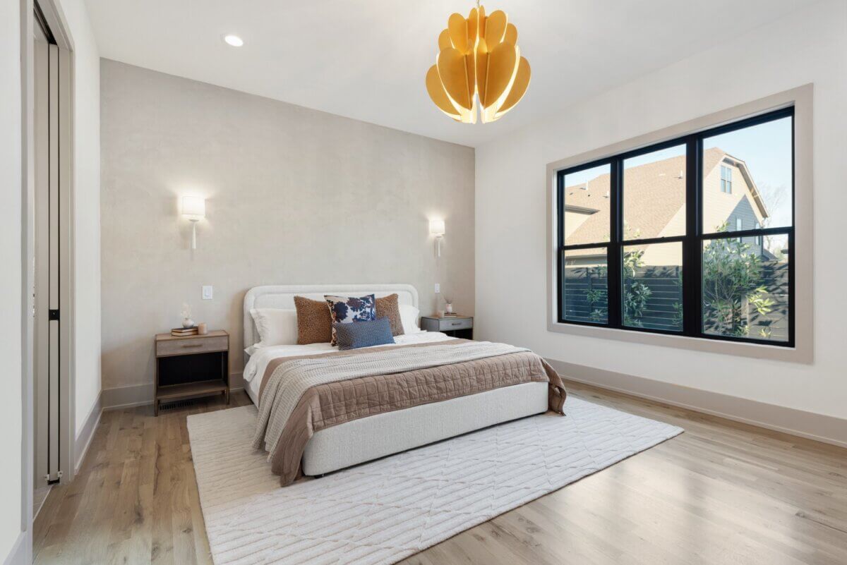 A Look Inside Brentwood's Newest Luxury Address - 8 Modern bedroom with a bed, nightstands, a large window, wooden floors, neutral decor, and a yellow pendant light fixture hanging from the ceiling—an inviting retreat often featured in Nashville luxury homes for sale.
