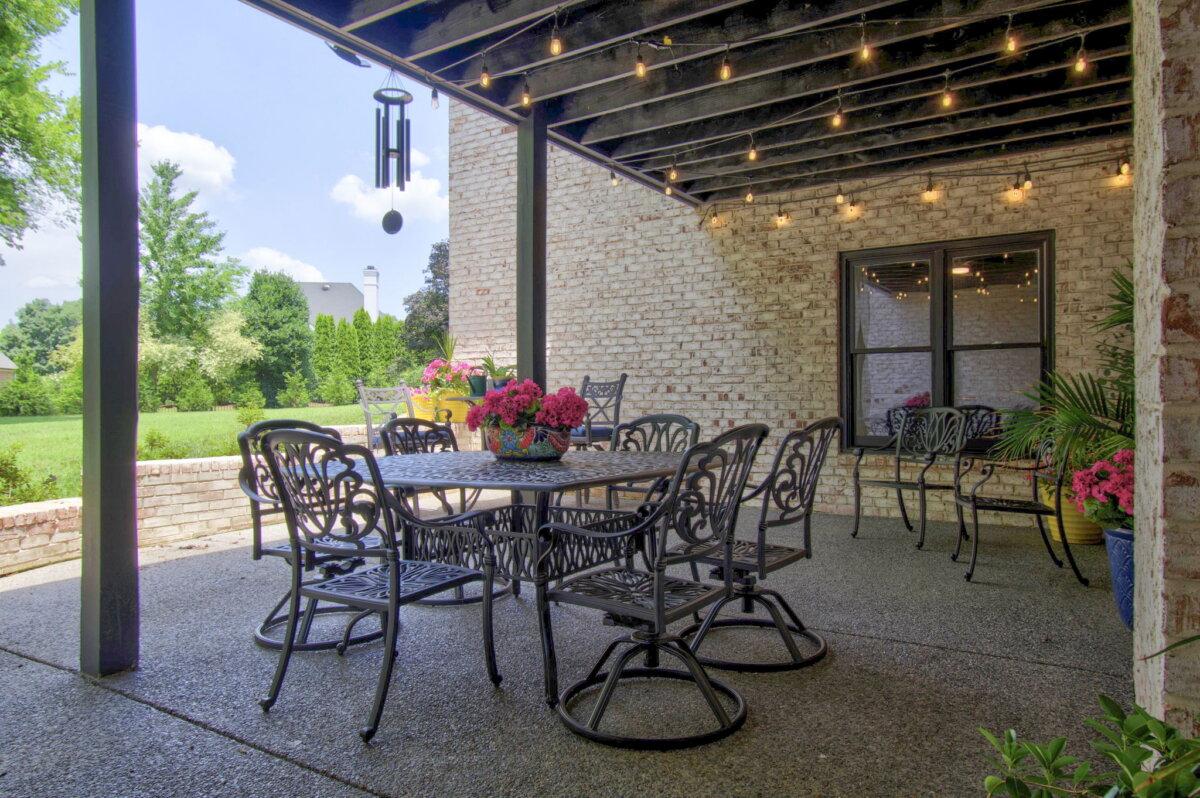 Just Listed! This Renovated Brentwood Home Has a Private Primary Wing, 4 Fireplaces & a Park-Like Backyard - 19 Enjoy a covered patio in this renovated Brentwood home, featuring metal chairs and tables, string lights overhead, potted flowers on tables, and a park-like backyard with views of a green lawn and trees in the background.