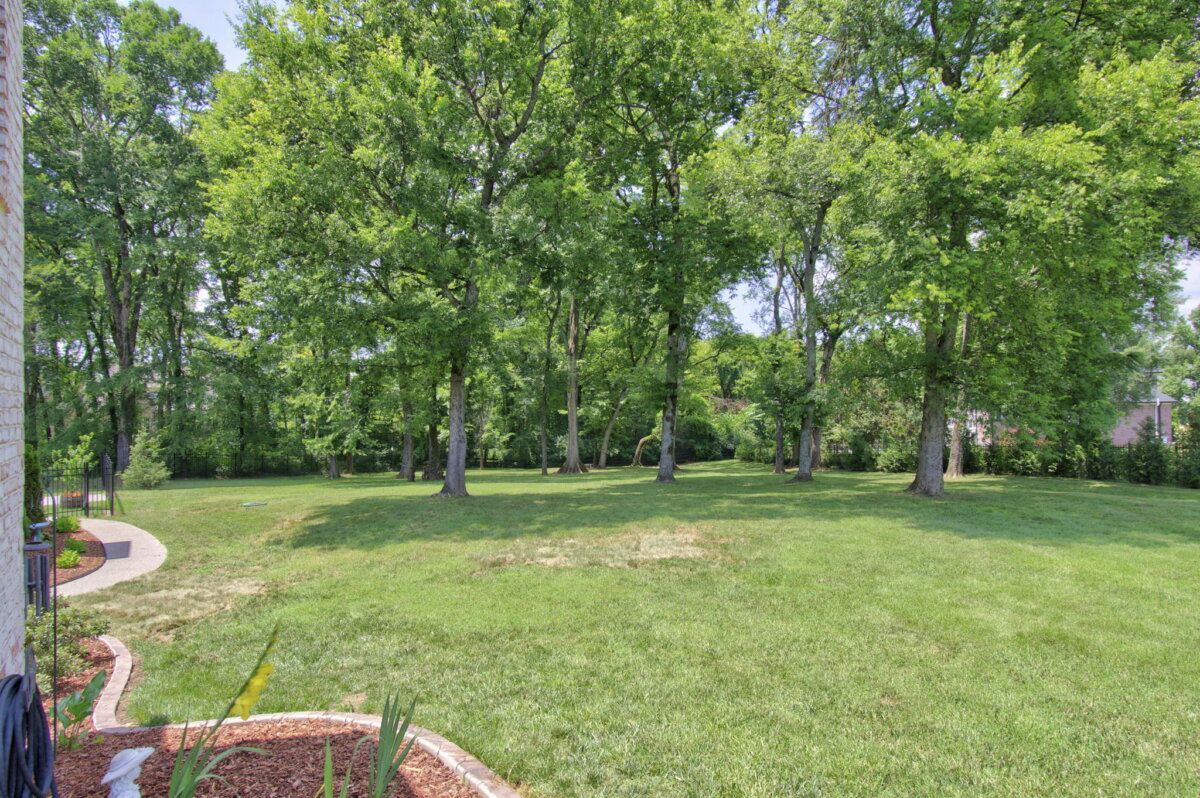 Just Listed! This Renovated Brentwood Home Has a Private Primary Wing, 4 Fireplaces & a Park-Like Backyard - 18 A spacious, park-like backyard with green grass, several tall trees, and a curved walkway along the left side bordered by landscaped plants, offering a tranquil retreat behind this beautifully renovated Brentwood home.
