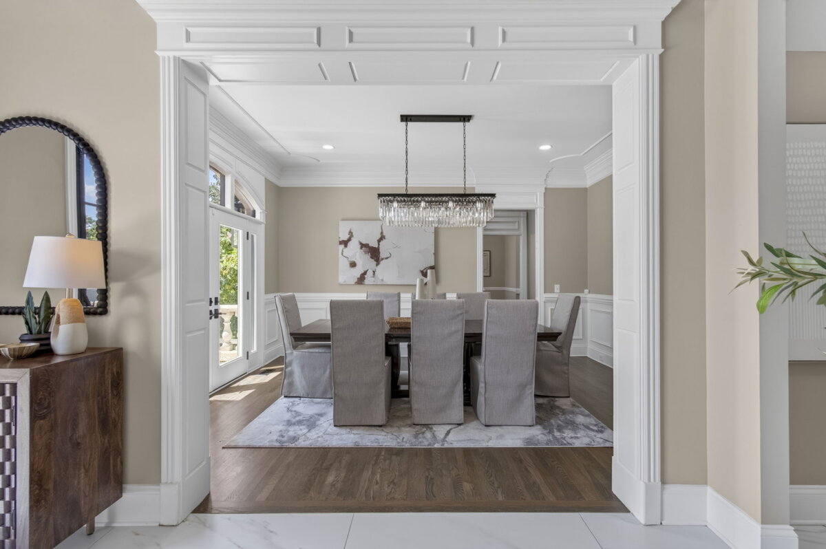 Just Listed! This Renovated Brentwood Home Has a Private Primary Wing, 4 Fireplaces & a Park-Like Backyard - 6 A formal dining room with a long table, eight upholstered chairs, a modern chandelier, wall art, and neutral decor. Large windows provide natural light and offer scenic views of the park-like backyard in this beautifully renovated home.