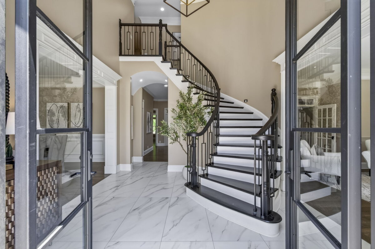 Just Listed! This Renovated Brentwood Home Has a Private Primary Wing, 4 Fireplaces & a Park-Like Backyard - 4 Open double doors reveal a spacious entryway with a curved staircase with black railings, beige walls, and a hallway leading to other rooms in this beautifully renovated Brentwood home.