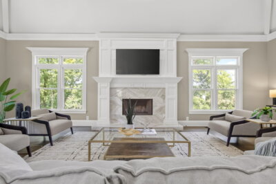 Elegant living room with beige walls, large windows, a marble fireplace, a wall-mounted TV, neutral armchairs, and a glass coffee table with decor in this beautifully renovated Brentwood Home overlooking a park-like backyard.
