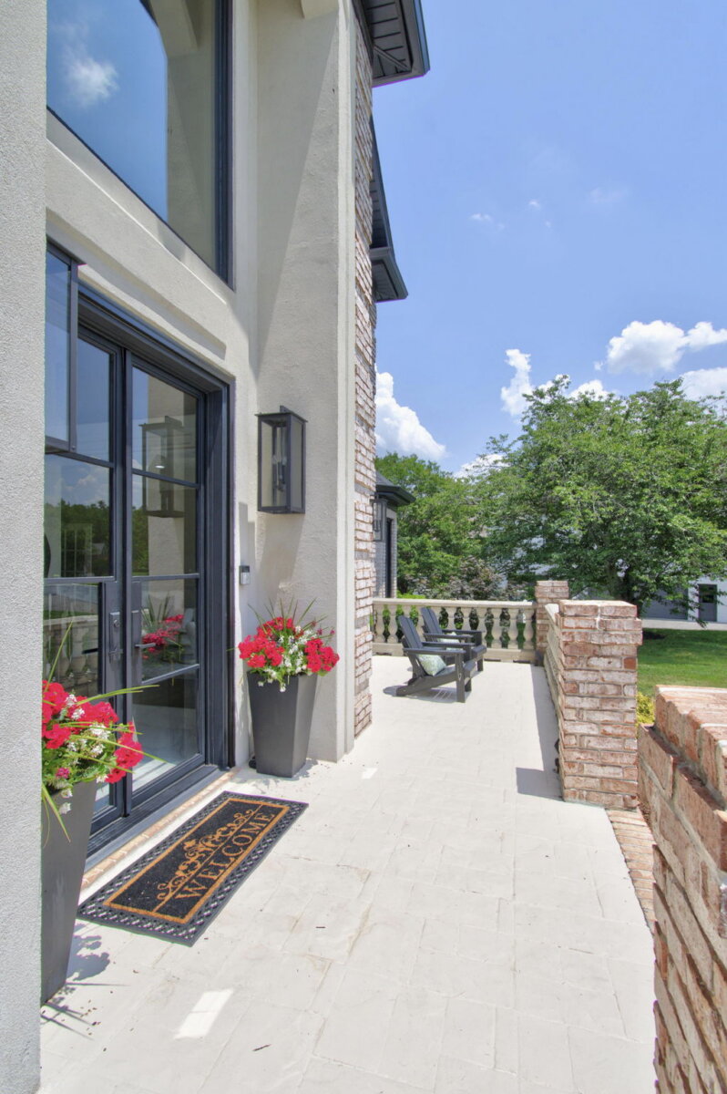 Just Listed! This Renovated Brentwood Home Has a Private Primary Wing, 4 Fireplaces & a Park-Like Backyard - 3 Modern patio with glass doors, two flower planters, a welcome mat, and brick walls. Two chairs face a park-like backyard under a blue sky with scattered clouds, creating a tranquil atmosphere perfect for this renovated Brentwood home.
