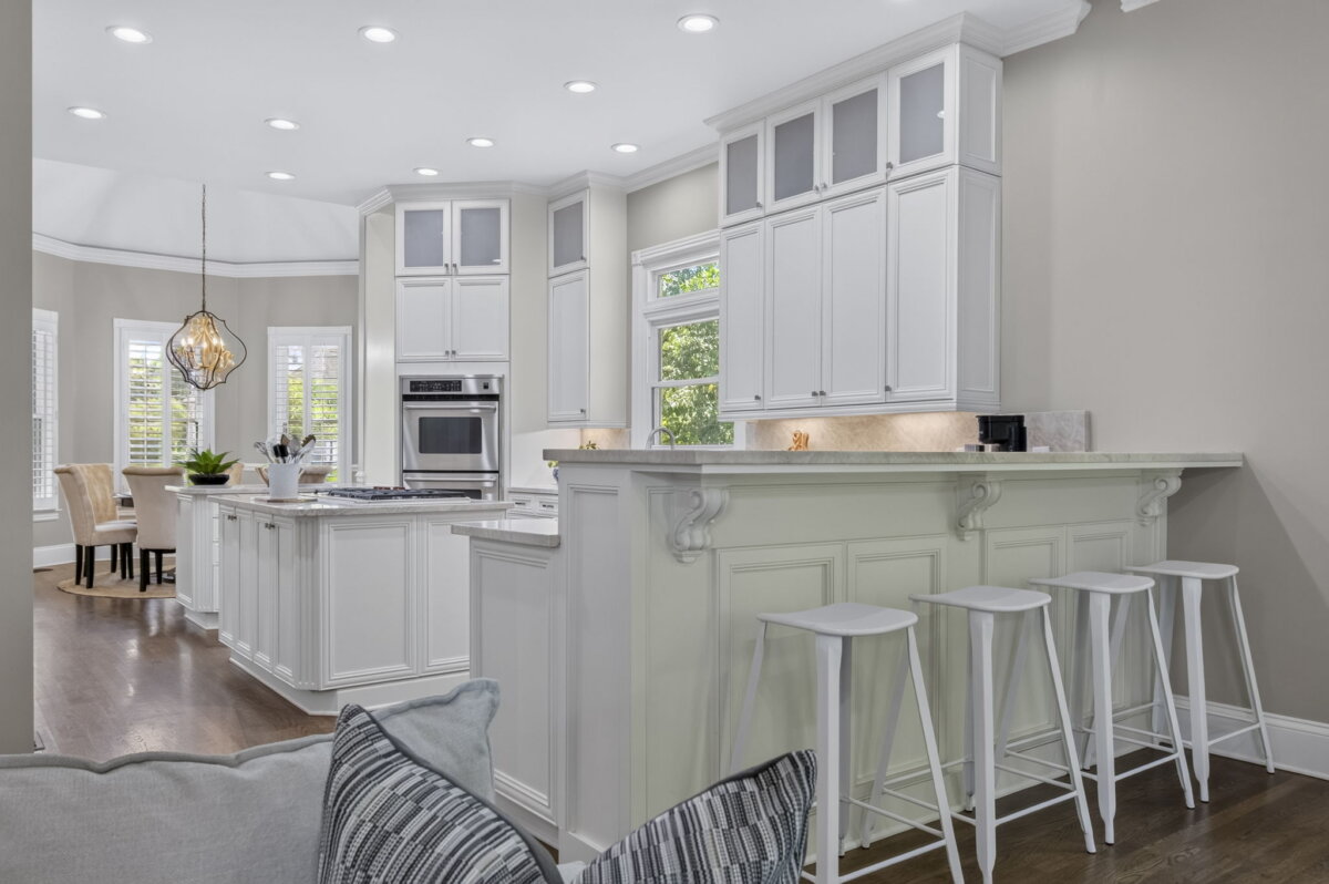 Just Listed! This Renovated Brentwood Home Has a Private Primary Wing, 4 Fireplaces & a Park-Like Backyard - 10 Modern kitchen with white cabinets, bar seating with three stools, stainless steel appliances, light countertops, and a view of a dining area with a chandelier and windows in the background. This beautifully renovated Brentwood Home also offers seamless access to a park-like backyard for effortless indoor-outdoor living.