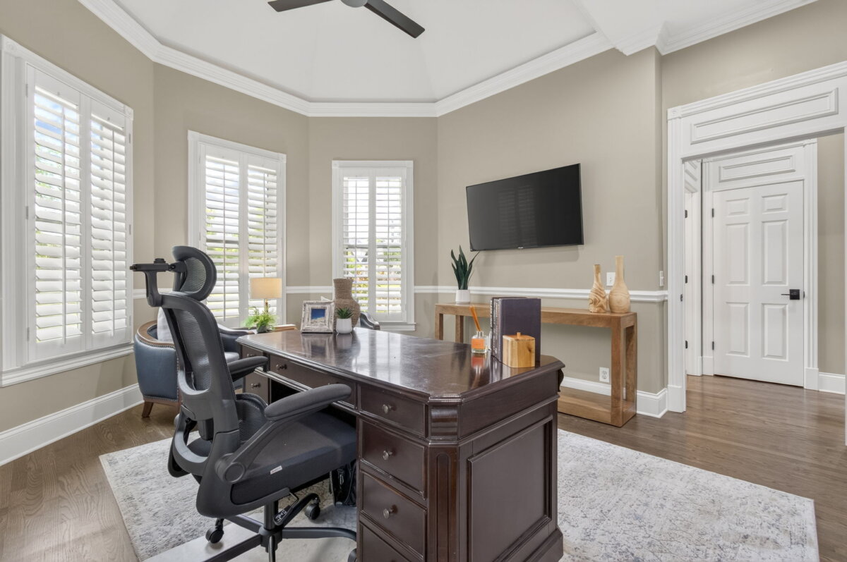Just Listed! This Renovated Brentwood Home Has a Private Primary Wing, 4 Fireplaces & a Park-Like Backyard - 14 Home office with a dark wooden desk, office chair, wall-mounted TV, console table with decor, large windows with shutters, and hardwood floorsβperfectly complementing this renovated Brentwood home.